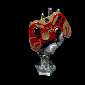 May include: A red and gold video game controller is held by a silver robotic hand. The controller is on a silver stand with a mechanical design.