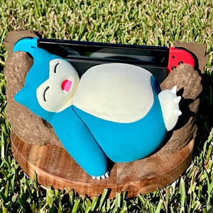 May include: A blue and white Snorlax figure rests on a wooden base, cradling a Nintendo Switch. The figure has a light blue body, white belly, and a pink mouth. The Switch has blue and red controllers.