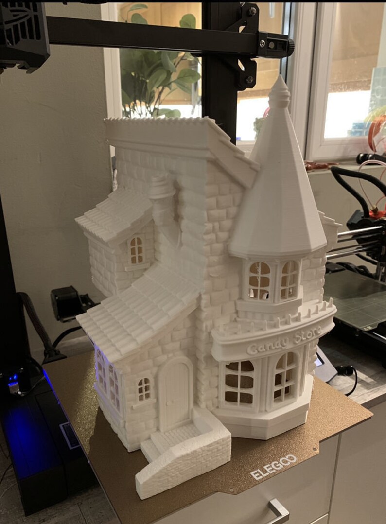 May include: A white 3D printed model of a small house with a candy store sign that reads "Candy Store". The house has a pointed roof, a chimney, and a small porch with steps. The model is sitting on a brown surface.