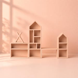 May include: A set of light pink wooden shelves in the shape of houses and a teepee. The shelves have various compartments and are arranged on a matching pink surface. The design is minimalist and modern.