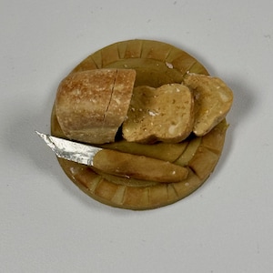 May include: A miniature bread loaf, partially sliced, with two slices and a small knife on a round wooden cutting board. The bread is light brown with a textured surface. The knife has a silver blade and a brown handle. A food-themed miniature.