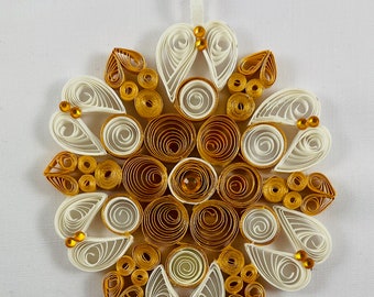 May include: A handcrafted, circular ornament with a detailed quilling design. The ornament features a combination of white, gold, and brown paper, arranged in a symmetrical pattern. Small gold beads are used as accents. A white ribbon is attached for hanging.