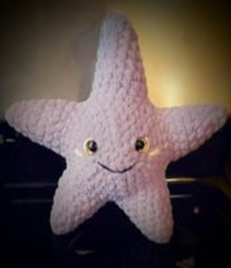 May include: A plush, lavender-colored starfish with a smiling face and small, black eyes. The five-pointed star is made of a soft, textured material. The starfish is a handmade item.