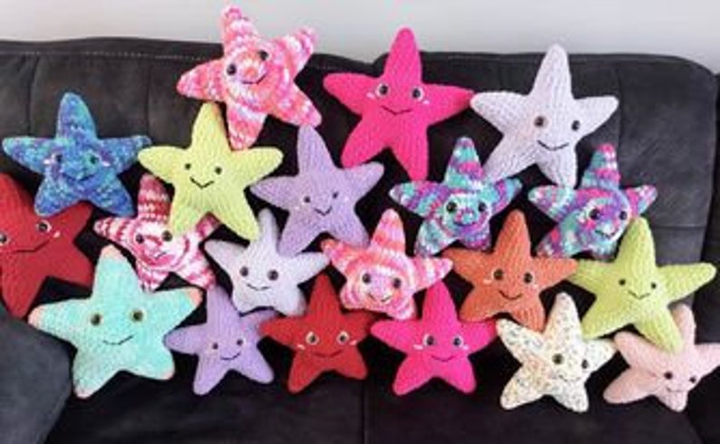 May include: A collection of colorful, crocheted star-shaped plush toys with smiling faces. The stars vary in color, including red, pink, blue, green, yellow, and purple. Each star has embroidered facial features and a soft, textured appearance.