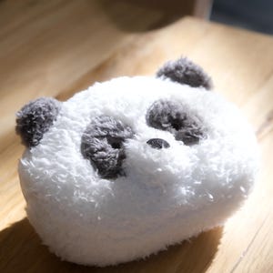 Puede incluir: Una cara de panda de peluche con pelaje blanco y detalles grises. El panda tiene orejas grises, parches oculares y una pequeña nariz negra. El juguete de peluche está sobre una superficie de madera.