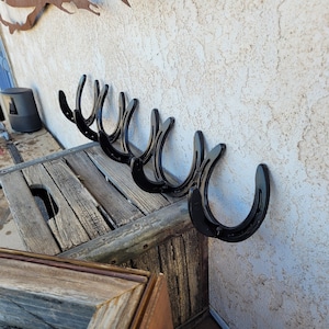 May include: A row of seven black metal horseshoes mounted on a wooden wall, with a rustic wooden shelf below.