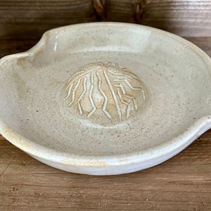 May include: A white ceramic citrus juicer with a textured surface and a raised, patterned center. The juicer has a spout for pouring juice.