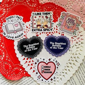 Pode incluir: Um conjunto de dez adesivos coloridos com vários desenhos e textos. Os adesivos apresentam corações, livros e textos como "Oops I Fell in Love Again", "The Stupidest Holiday of the Year", e "I Like Them Extra Spicy".