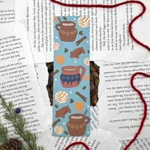May include: A blue and brown bookmark with illustrations of a mug of hot chocolate, cinnamon sticks, and Mexican pastries. The bookmark has a white background with snowflakes and stars.