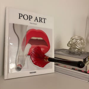 May include: A hardcover book titled POP ART by Klaus Honnef published by TASCHEN. The cover features a graphic illustration of bright red lips and a hand holding a cigarette, with a black-handled makeup brush resting horizontally across the front.