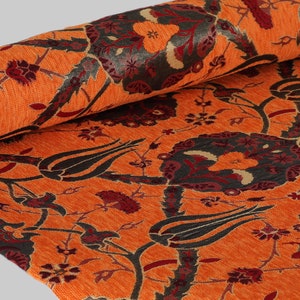 Op de afbeelding: Oranje stof met een herhalend patroon van gestileerde bloemen en bladeren in tinten zwart, rood en beige.