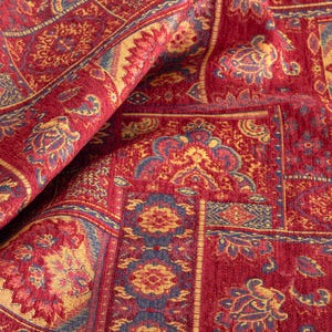 Könnte beinhalten: Nahaufnahme eines gefalteten roten Stoffes mit einem komplizierten Muster. Das Design zeigt florale und Paisley-Motive in Gold-, Blau- und Rottönen. Der Stoff hat ein strukturiertes Aussehen, was auf eine gewebte oder gestickte Konstruktion hindeutet.