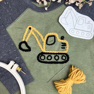 May include: A long-sleeve shirt with a sage green body and dark gray raglan sleeves, featuring a yellow and black embroidered excavator design. The shirt is accompanied by a white paper stencil of an excavator, a white embroidery hoop, and a needle with yellow yarn.