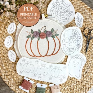 May include: An embroidery hoop containing a stitched pumpkin with floral accents, surrounded by various stick and stitch templates featuring a spiderweb, skull, ghost, bats, and pumpkins, along with three spools of embroidery thread and scissors.