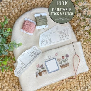 Könnte beinhalten: Eine cremefarbene Mütze mit Stickerei und einer Nadel mit rosa Faden. Die Stickerei zeigt Topfblumen, einen Schmetterling und einen leeren Rahmen. Zwei Garnrollen und eine PDF-Vorlage zum Aufkleben und Sticken sind ebenfalls sichtbar.