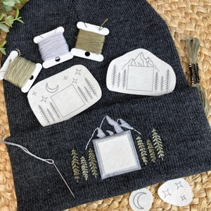 May include: Dark gray beanie with embroidered mountain and tree designs. Includes spools of thread and patches with mountain, moon, and star motifs. A needle and thread are positioned near the embroidered design, suggesting a DIY craft project.