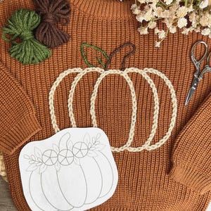 May include: A rust-coloured knit jumper featuring a large embroidered pumpkin design in cream yarn with green and brown stem details. The jumper is accompanied by two small yarn bundles, a pair of silver scissors, and a white paper pumpkin stencil.