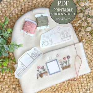 Puede incluir: Un gorro de punto color crema con diseños florales y de jardín bordados, acompañado de dos plantillas de papel blanco para bordar, dos pequeños carretes de hilo rosa y verde oliva, y una aguja de coser.