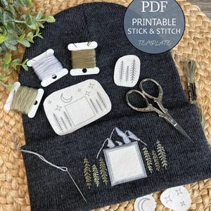 Puede incluir: Un gorro gris oscuro con suministros de bordado, que incluyen carretes de hilo, tijeras y una aguja. El gorro presenta diseños bordados de montañas y árboles, con el texto "PDF Printable Stick & Stitch Template."
