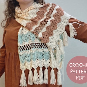 Peut inclure: Une écharpe en crochet avec un motif de couleur marron, blanc et bleu. L'écharpe a des franges en bas et est réalisée avec un point délicat. L'écharpe est drapé sur l'épaule d'une personne.