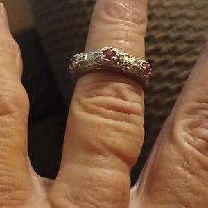 Può includere: Un anello in metallo color argento caratterizzato da un intricato motivo floreale o a viticcio testurizzato sulla fascia. L'anello è incastonato con tre piccole pietre rotonde rosso scuro distanziate uniformemente sulla superficie superiore.