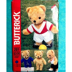 May include: A Butterick 201 craft sewing pattern envelope featuring a tan teddy bear wearing a red and white sports jersey and shorts with a yellow ball. Additional images show teddy bears in blue and white outfits on a green grass background.