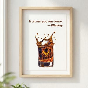 May include: A framed print featuring a watercolor illustration of a whiskey cocktail with ice, mid-splash. The artwork includes the quote, "Trust me, you can dance. -Whiskey" in a brown font. The frame is light wood.