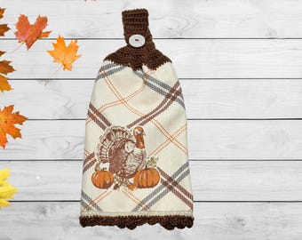May include: A hanging kitchen towel featuring a cream-colored fabric with a brown and orange plaid pattern. The towel displays a graphic of a turkey and two pumpkins. It has a dark brown crocheted top with a white button and a matching crocheted edge.