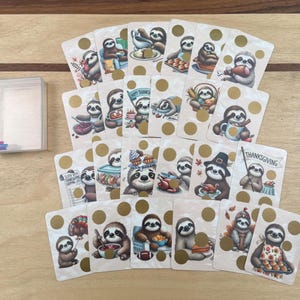 May include: A collection of illustrated cards featuring sloths in various Thanksgiving-themed scenes. Each card has a gold polka dot and a sloth preparing food or holding a sign that says "Thanksgiving". A clear plastic card holder is on the left.