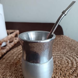 Peut inclure: Une calebasse à maté argentée avec une paille en métal, posée sur un tapis tissé. La calebasse a un design bicolore, avec une partie supérieure polie et une partie inférieure mate. La paille est incurvée et possède un filtre. Hauteur : environ 8 cm.