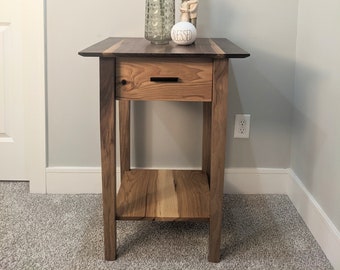 May include: A wooden end table with a single drawer and a lower shelf. The table is made of walnut and oak wood and has a natural finish. A vase with dried pampas grass and a small ceramic figurine are on top of the table.