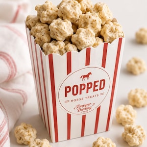 Puede incluir: Un recipiente de cartón con rayas rojas y blancas lleno de golosinas para caballos de color tostado claro y forma de estrella. El recipiente presenta un logotipo circular con una silueta de caballo, el texto POPPED HORSE TREATS y la marca Dapper Darling.
