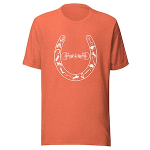 Puede incluir: Camiseta naranja con un diseño de herradura blanca. La herradura está llena de varios símbolos ecuestres, incluyendo un estribo, una fusta y una cabeza de caballo.
