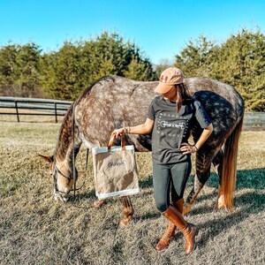 May include: A woman wearing a black t-shirt with the text "Road Less Traveled" and a white graphic of a horse bit, stands next to a brown and white horse in a field. She is holding a brown and white canvas tote bag with the word "Ghost" printed on it.