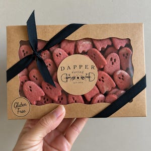 Könnte beinhalten: Eine rechteckige braune Pappschachtel mit Sichtfenster, gefüllt mit roten, geisterförmigen Keksen. Die Schachtel ist mit einem schwarzen Band umwickelt und mit einem runden Aufkleber DAPPER darling EST 2018 sowie einem Gluten Free Etikett versehen.