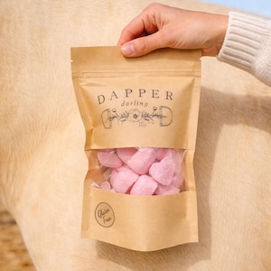 May include: A brown paper bag with a clear window, filled with pink heart-shaped candies. The bag has the words "Dapper Darling" printed on it, along with a floral design and the words "Gluten Free".
