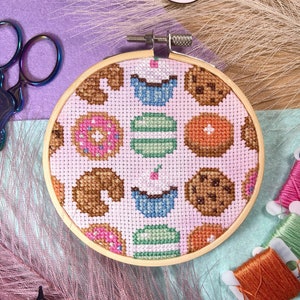 May include: A pink and white cross-stitch embroidery hoop with a design of donuts, cupcakes, and cookies. The hoop is made of wood and has a brown finish.