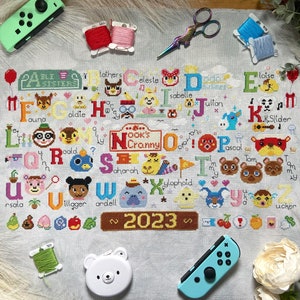 May include: A cross-stitch sampler featuring the alphabet with characters from the Animal Crossing video game. The design includes the text "Nook's Cranny" and "2023". The image is surrounded by craft supplies and a Nintendo Switch controller.
