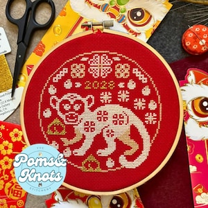 May include: A red embroidery hoop with a white monkey design and gold accents. The design includes the year 2022 and the text "Poms & Knots Stitchery".