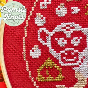 以下が含まれることがあります： 白と金のデザインが施された赤いクロスステッチ刺繍枠。デザインは、金色のセンターを持つスタイリッシュな花柄です。枠には「Poms & Knots Stitchery」とラベルが貼られています。