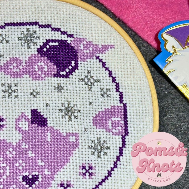 May include: A cross-stitch embroidery in a wooden hoop, featuring a purple and light purple design with silver snowflakes. The design is on white fabric. A pink felt square and a decorative pin are in the background. The logo for Poms & Knots Stitchery is in the lower right.