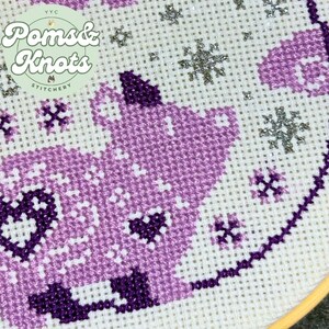 May include: Close-up of a cross-stitch design featuring a purple cat with heart details and snowflake accents. The design is on white fabric and includes the text "Poms & Knots Stitchery".