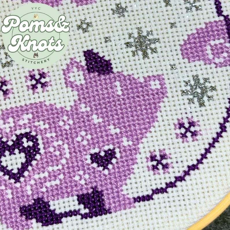 May include: Close-up of a cross-stitch design featuring a purple cat with heart details and snowflake accents. The design is on white fabric and includes the text "Poms & Knots Stitchery".