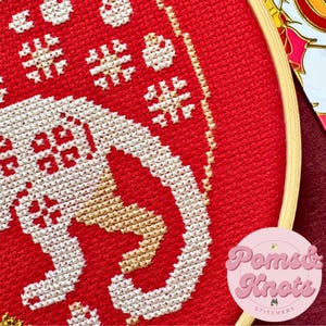 以下が含まれることがあります： 白と金の象のデザインが施された赤いクロスステッチ刺繍枠。枠は木製のフレームで囲まれています。デザインには雪片やその他の幾何学模様が含まれています。右下隅には「Poms & Knots Stitchery」の文字が見えます。