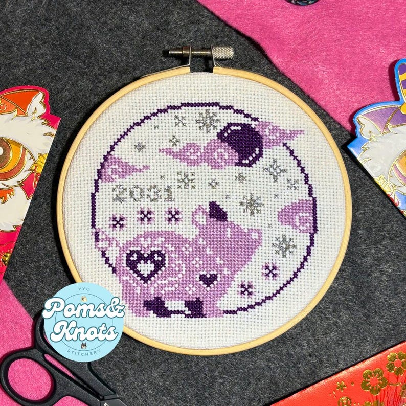 May include: A round cross-stitch embroidery hoop featuring a purple pig design with a heart and the year 2021. The design includes snowflakes and a moon. The hoop is set against a dark gray background.