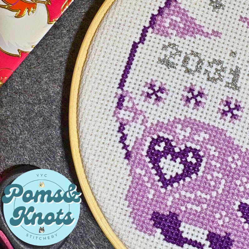 May include: A cross-stitch embroidery hoop with a purple and white design. The design includes the number "2023" and floral patterns. The hoop is made of wood and the fabric is white. The logo "Poms & Knots Stitchery" is visible.
