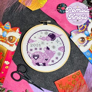May include: A cross-stitch embroidery hoop featuring a purple and white design with the year 2031. The design includes a pig, clouds, and stars. The image also includes red envelopes, scissors, and a pink felt background.