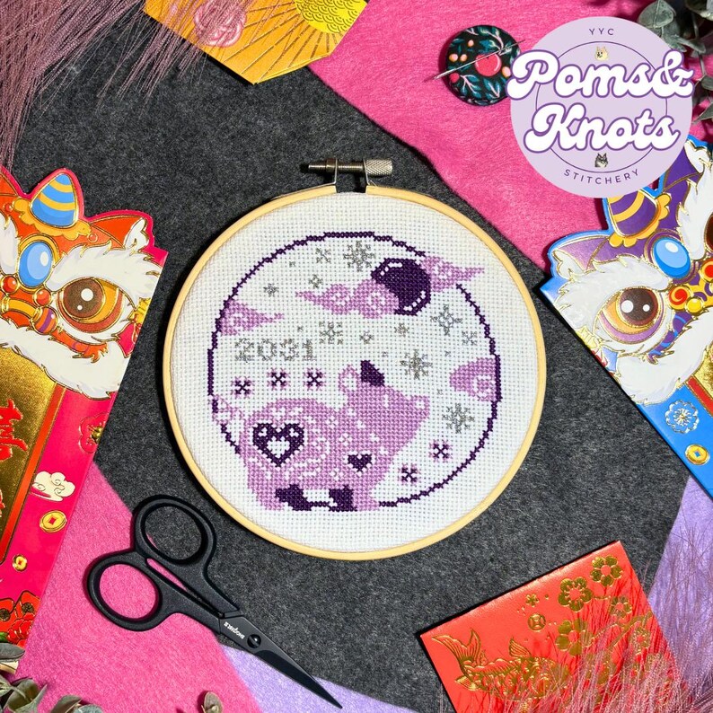 May include: A cross-stitch embroidery hoop featuring a purple and white design with the year 2031. The design includes a pig, clouds, and stars. The image also includes red envelopes, scissors, and a pink felt background.