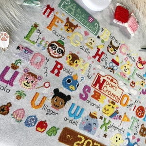May include: A cross-stitch sampler featuring the alphabet with characters from the Animal Crossing video game. Each letter is accompanied by a character's name and image. The sampler includes the text "Nook's Cranny" and the year "2023".