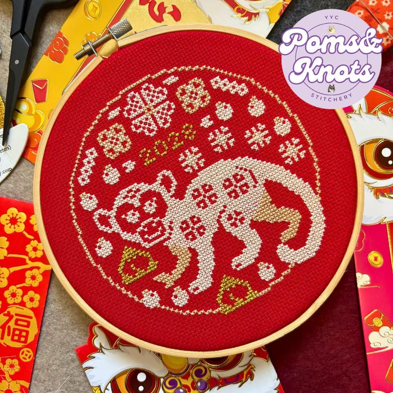 以下が含まれることがあります： 赤い刺繍枠に、白いクロスステッチで猿と2022年のデザインが施されています。デザインは、金色と白のクロスステッチのシンボルで囲まれています。枠には「Poms & Knots Stitchery」とラベルが貼られています。